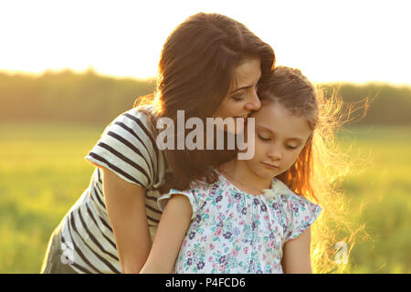 Emotionale ruhe Mutter Sie entspannende kid Mädchen mit liebenden Augen auf Sonnenuntergang hellen Sommer Hintergrund umarmte. Closeup helle Farbe portrait Stockfoto