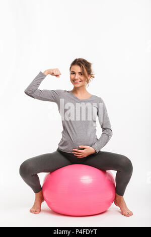 Glücklich schwanger Frau sitzt auf Fitness Ball und zeigt Muskeln isoliert auf weißem Stockfoto