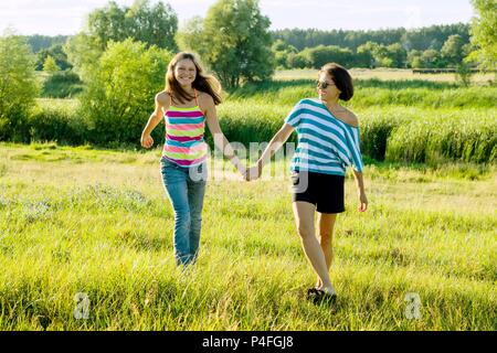 Eltern und Teenager, glückliche Mutter und jugendlich Tochter 13, 14 Jahre alt, halten die Hände gehen, lachen, sprechen. Hintergrund Natur, Park Stockfoto