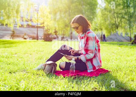 Erwachsene im mittleren Alter Frau sitzt im City Park auf dem Gras. Über Kopfhörer hört, Hörbuch, Uhren Video, Selbst-Bildung Stockfoto