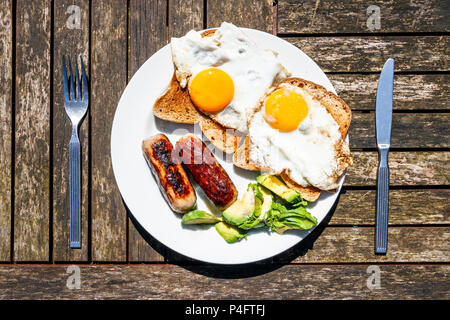 Weiße Platte mit Spiegelei auf Toast, Würstchen und Avocado mit Metall Messer und Gabel auf einem hölzernen Lattenrost Garten Tisch Stockfoto