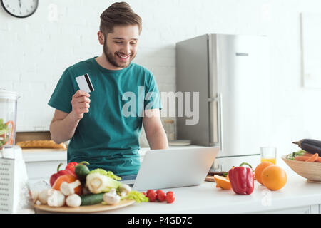Lächelnder Mann mit Kreditkarte und Laptop Kauf von Waren online in der Küche zu Hause. Stockfoto
