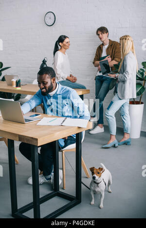 Lächelnd afrikanischen amerikanischen Geschäftsmanns, der auf dem Laptop und Halten der Hund an der Leine und Mitarbeiter in Sitzung hinter Stockfoto