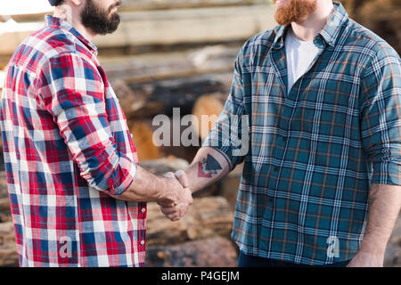 7/8 Schuß von zwei bärtigen Holzfäller in karierten Hemden Hände schütteln im Sägewerk Stockfoto