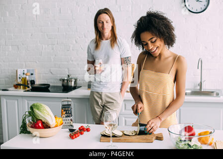 Lächelnd afrikanische amerikanische junge Frau die Aubergine und die Unterhaltung mit Wein Glas in der Küche zu Freund Stockfoto