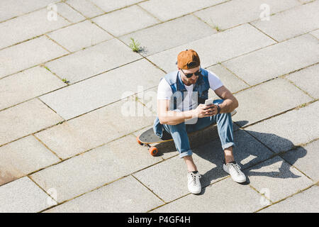 Hohe Betrachtungswinkel von hübscher junger Mann sitzt auf Longboard mit Smartphone Stockfoto