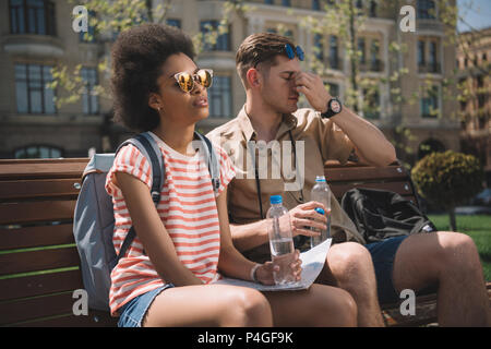 Müde interracial Paare von Reisenden mit Wasserflaschen und Karte sitzt auf der Bank Stockfoto