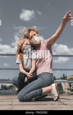 Mutter zeigt auf etwas Tochter in Schutzmaske auf Brücke, Luftverschmutzung Konzept Stockfoto