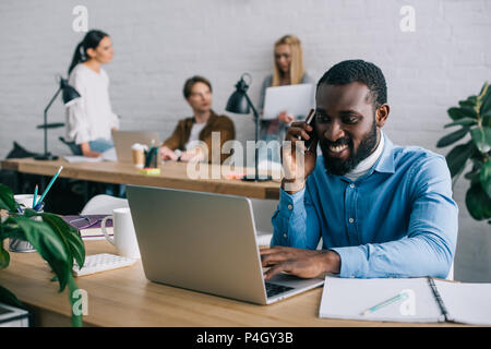 Afrikanische amerikanische Geschäftsmann sprechen auf Smartphone und mit Laptop am Tisch und Kollegen hinter arbeiten Stockfoto