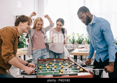 Seitenansicht der glücklichen multikulturellen Unternehmer Spielen Tischfußball vor Feiern und gestikulierend weiblichen Kollegen Stockfoto