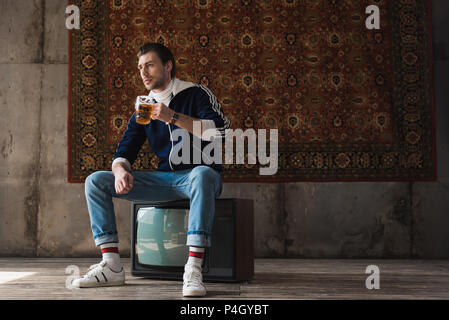 Nachdenklicher junger Mann in Vintage Kleidung mit Becher Bier sitzen auf Retro tv an der Decke hängend an der Wand Stockfoto