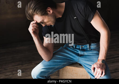Nachdenklichen jungen Mann im schwarzen T-Shirt und Hosenträger sitzen auf hölzernen Kasten Stockfoto