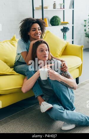 Attraktive afrikanische amerikanische Frau Fernsehen und umarmt Freund mit tätowierten Hand zeigen auf TV-Bildschirm Stockfoto