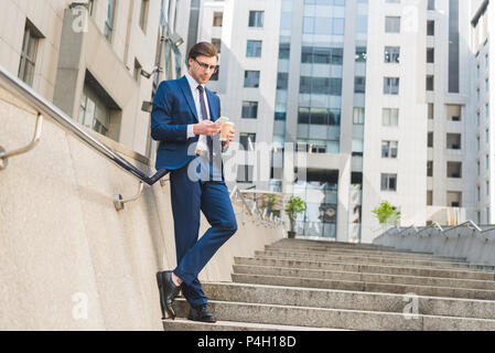 Attraktive junge Unternehmer im eleganten Anzug mit Papier Tasse Kaffee mit Smartphone auf der Treppe in der Nähe von Business Gebäude Stockfoto