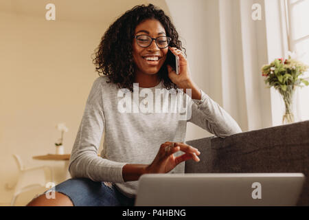 Geschäftsfrau auf dem Sofa zu Hause sitzt und spricht über Handy während der Arbeit am Laptop. Lächelnde Frau in modischen zerrissenen Jeans Arbeiten am Laptop Stockfoto