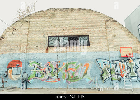 Rückansicht des Menschen malen bunte Graffiti an der Wand mit Basketballkorb und Leitern Stockfoto