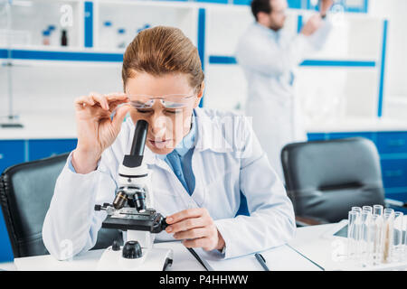 Selektiver Fokus der Wissenschaftlerin durch Mikroskop auf Reagenz am Arbeitsplatz mit Kollegen hinter im Labor Stockfoto
