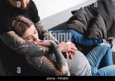 Zugeschnittenes Bild von tätowierten Freund umarmen schwangere Freundin auf dem Sofa zu Hause Stockfoto
