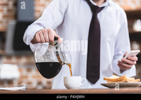 Zugeschnittenes Bild der Einzelgänger Geschäftsmann ausgießen und Smartphone Holding in Küche Stockfoto