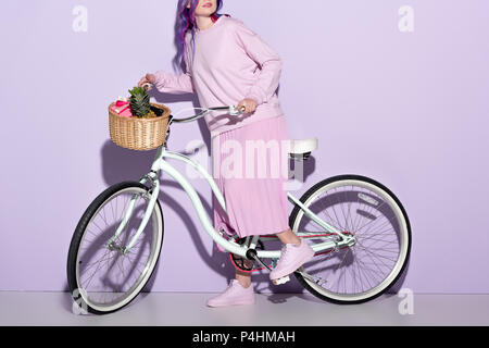 7/8 shot der jungen Frau in rosa Kleidung auf dem Fahrrad mit Ananas und Bananen im Korb Stockfoto