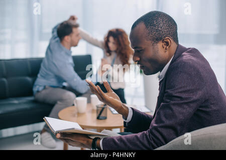 African American Ratgeber suchen in Lehrbuch während scheidene Paare kämpfen auf dem Sofa Stören Stockfoto