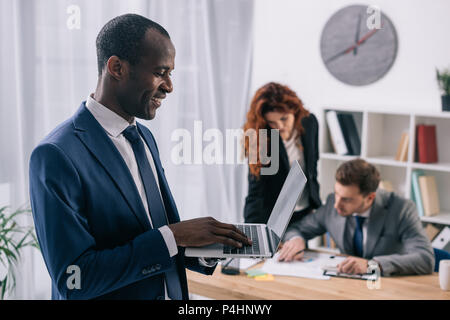 Lächelnd afrikanischen Geschäftsmann mit Laptop in den Händen und zwei Kollegen am Tisch arbeiten Stockfoto