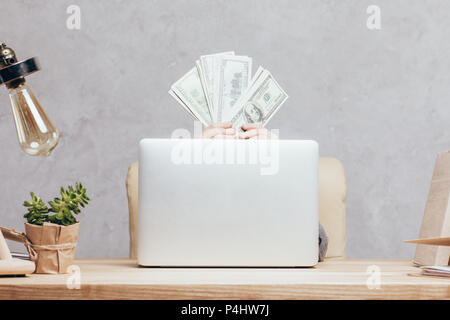 Teilweise mit Blick auf die Kind mit Geld in den Händen versteckt sich hinter Laptop am Arbeitsplatz isoliert auf Grau Stockfoto