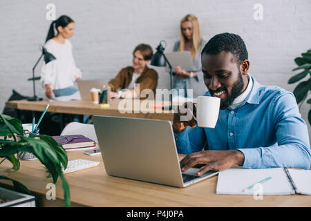 Lächelnd afrikanische amerikanische Geschäftsmann mit Laptop und trinken Kaffee aus der Tasse und Kollegen hinter der Arbeit in modernen Büro Stockfoto