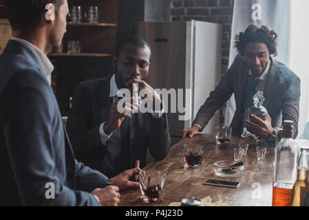 Drei multiethnischen Männer in Anzügen rauchen Zigarren und trinken Alkohol zusammen Stockfoto