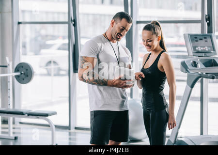 Lächelnden jungen Sportlerin durch die Finger, der auf Zeitplan auf digital Tablet in persönlichen Trainer im Fitnessraum Stockfoto