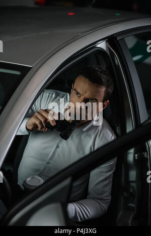 Hohe Betrachtungswinkel der männlichen Privatdetektiv mit Kaffee Tasse tun Überwachung durch Fernglas aus dem Auto Stockfoto