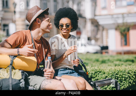 Happy interracial Paare der Touristen mit den Flaschen Wasser sitzt auf der Bank Stockfoto