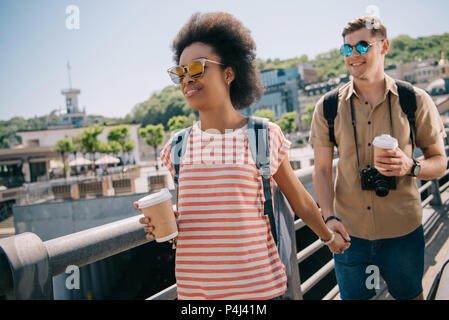 Interracial Paar Touristen mit Papier Tassen Kaffee und Kamera halten sich an den Händen und laufen auf Brücke Stockfoto