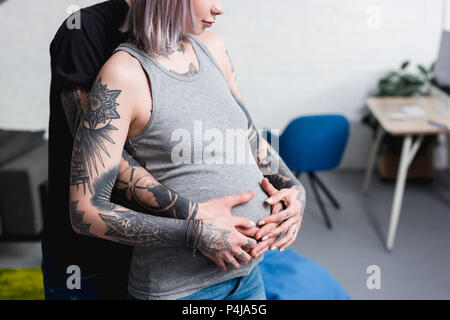 Zugeschnittenes Bild von tätowierten Freund umarmen schwangere Freundin aus Zurück im Wohnzimmer Stockfoto