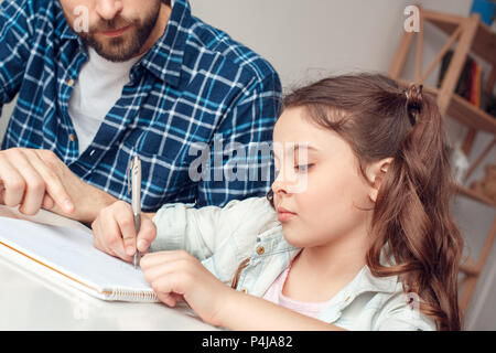 Bärtiger Mann und kleine Mädchen zu Hause Zeit mit der Familie am Tisch Tochter tun Übung schreiben am Notebook sitzen konzentriert close-up Stockfoto