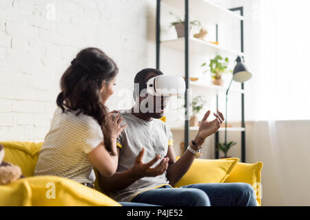 Junge afrikanische amerikanische Frau in der Nähe von schockiert Freund sitzen, während er das Tragen von virtual reality Headset auf der Couch zu Hause Stockfoto