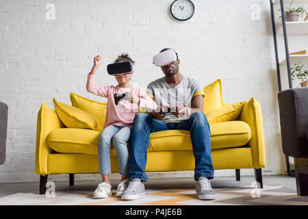 Junger Vater und Tochter im VR-Headsets spielen Videospiele auf der Couch zu Hause Stockfoto