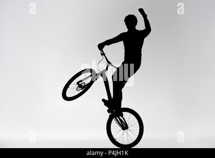 Silhouette des Versuches biker Stehen auf der Rückseite der Rad- und heben die Hand auf Weiß Stockfoto