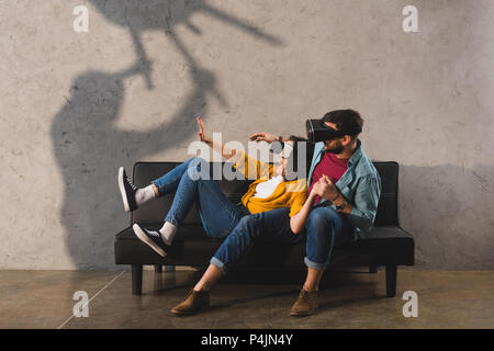 Unheimliche Schatten holding Stuhl und erschrocken Paar in virtual reality Headset auf der Couch Stockfoto