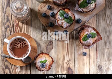 Muffins mit Heidelbeeren und eine Tasse heiße Schokolade auf einem hölzernen Hintergrund. Hausgemachte backen. Ansicht von oben Stockfoto