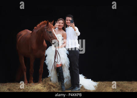 Junge jungvermählte Paar, gutaussehenden Bräutigam und schön schönheit Braut in modische Weiße Braut Hochzeit Kleid selfie Smartphone mit mit Pferd auf Schwarz Stockfoto