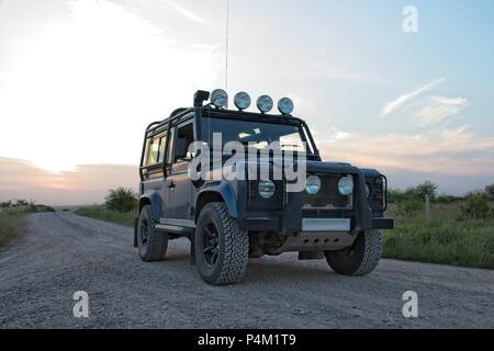 Eine blaue Offroad Land Rover Defender 90, Grafschaft auf grau Track mit Sonnenuntergang hinter. Stockfoto