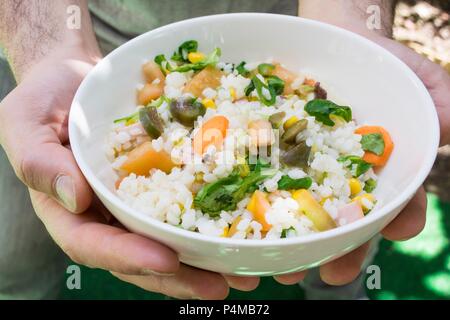 Hände halten eine Schüssel Reis Salat mit Tomaten, Basilikum, Olivenöl, Oliven, Mais und Karotten Stockfoto