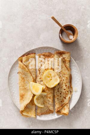 Drei gerollte Pfannkuchen pn ein Weiß Textur Platte mit zitronekeile auf einem steinernen Oberfläche propped mit hölzernen Sugar Bowl und Löffel Stockfoto