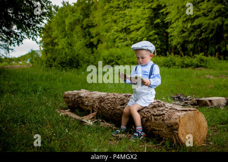 Ein litttle zwei Jahre alte Junge sitzt auf einem Baum und liest ein Buch. hildren Tag. Stockfoto