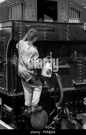 Portrait, schwarz-weiß Rückansicht des Dampfzugfahrers in schmutzigen Overalls, von hinten, Klettern an Bord Vintage UK Dampflokomotive Lampe zu wechseln. Stockfoto