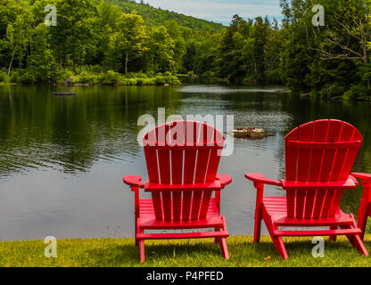 Zwei helle rote Adirondack Stühle am Ufer des See im Sommer mit einem schwimmenden Blumenkasten mit Blumen blühen Stockfoto