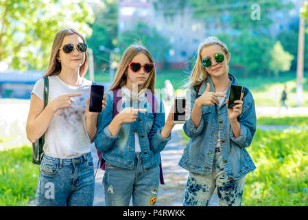 Drei Mädchen Schulmädchen. Sommer in der Natur. In den Händen hält. Geste Geste ist Smartphone angezeigt. In jeans Kleidung in Sonnenbrille. Konzept der Smartphone-anwendung. Emotionen interessante Gadget. Stockfoto