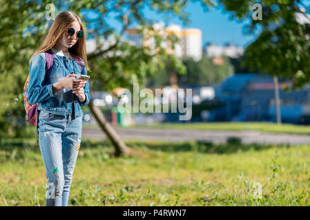 Kleines Mädchen Schulmädchen. Sommer im Park in der Natur nach der Schule. Schreibt ein Smartphone Nachricht. Freier Platz für Text. Stockfoto