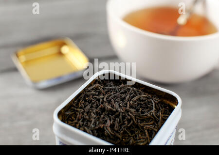 Schwarz loser Tee in Metalldose, mit unscharfen heiße Tasse gelb Drink im Hintergrund. Stockfoto